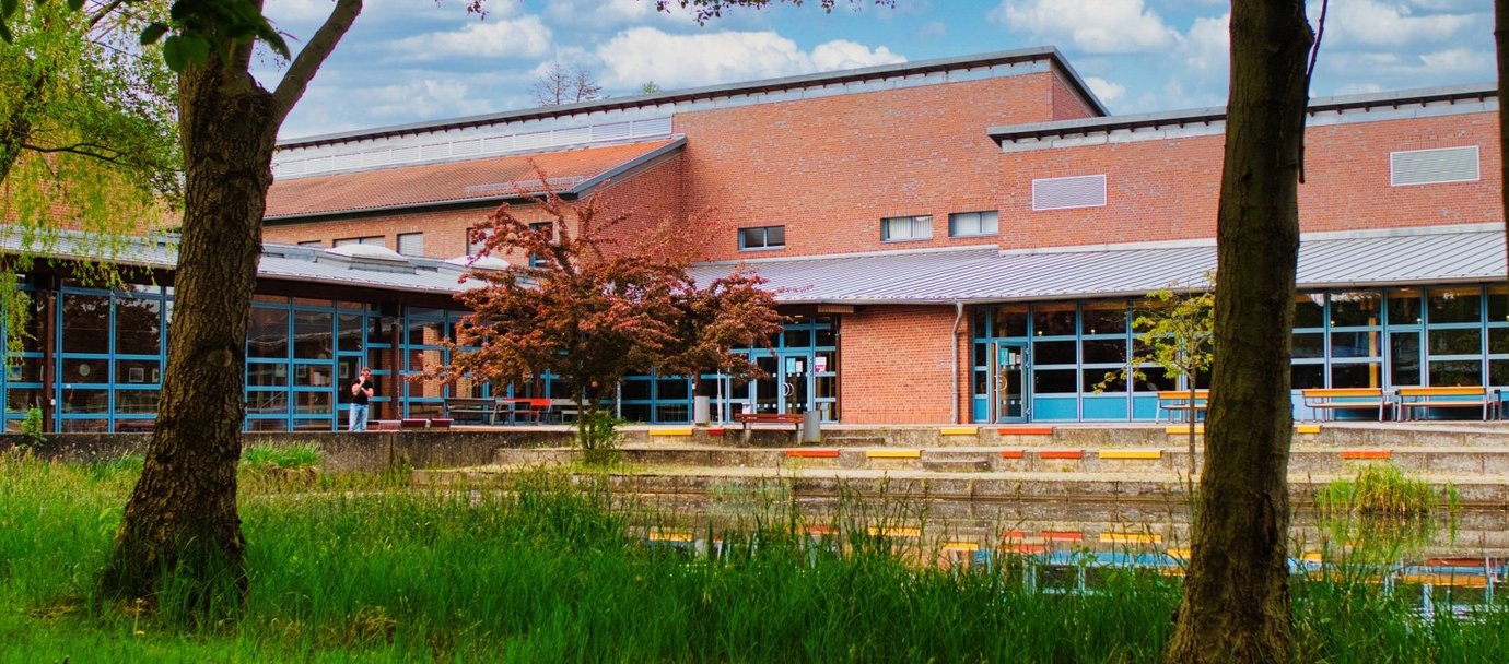 Bild vom Campus-Teich mit Bäumen im Vordergrund und Blick auf die Gebäude mit der Mensa im Hintergrund