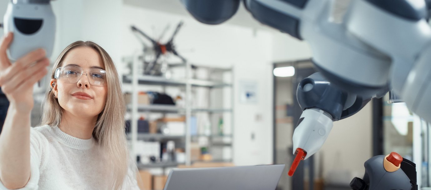 Studentin mit Schutzbrille steht vor einem Roboter und hält ihn mit einer Hand fest